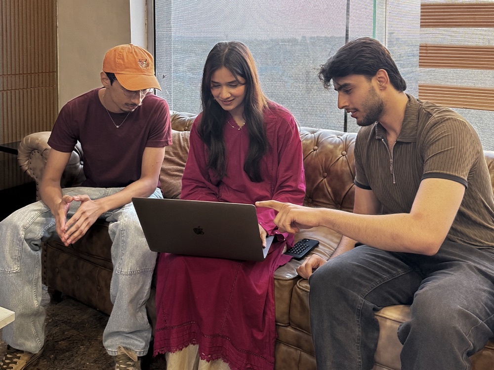 Three colleagues sitting together on a sofa around one open laptop, one of them pointing at the screen.